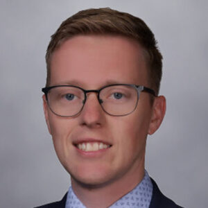 A young man with short light brown hair, wearing glasses, a navy suit, a light blue shirt, and a patterned tie, smiles at the camera against a plain gray background.
