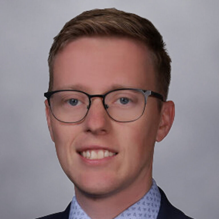 A young man with short light brown hair, wearing glasses, a navy suit, a light blue shirt, and a patterned tie, smiles at the camera against a plain gray background.