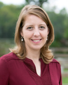 Woman with shoulder-length light brown hair, wearing a burgundy blouse and pearl earrings, smiling outdoors with green trees and a wooden fence blurred in the background.