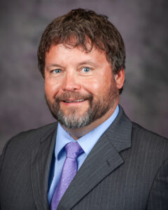 A man with brown hair and a beard is wearing a gray pinstripe suit, light blue shirt, and lavender tie, smiling in front of a blurred gray-purple background.