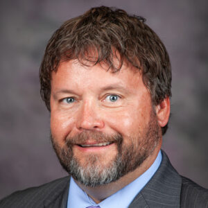 Man with wavy brown hair and beard, smiling, wearing a gray suit jacket, light blue shirt, and striped tie, in front of a blurred gray background.