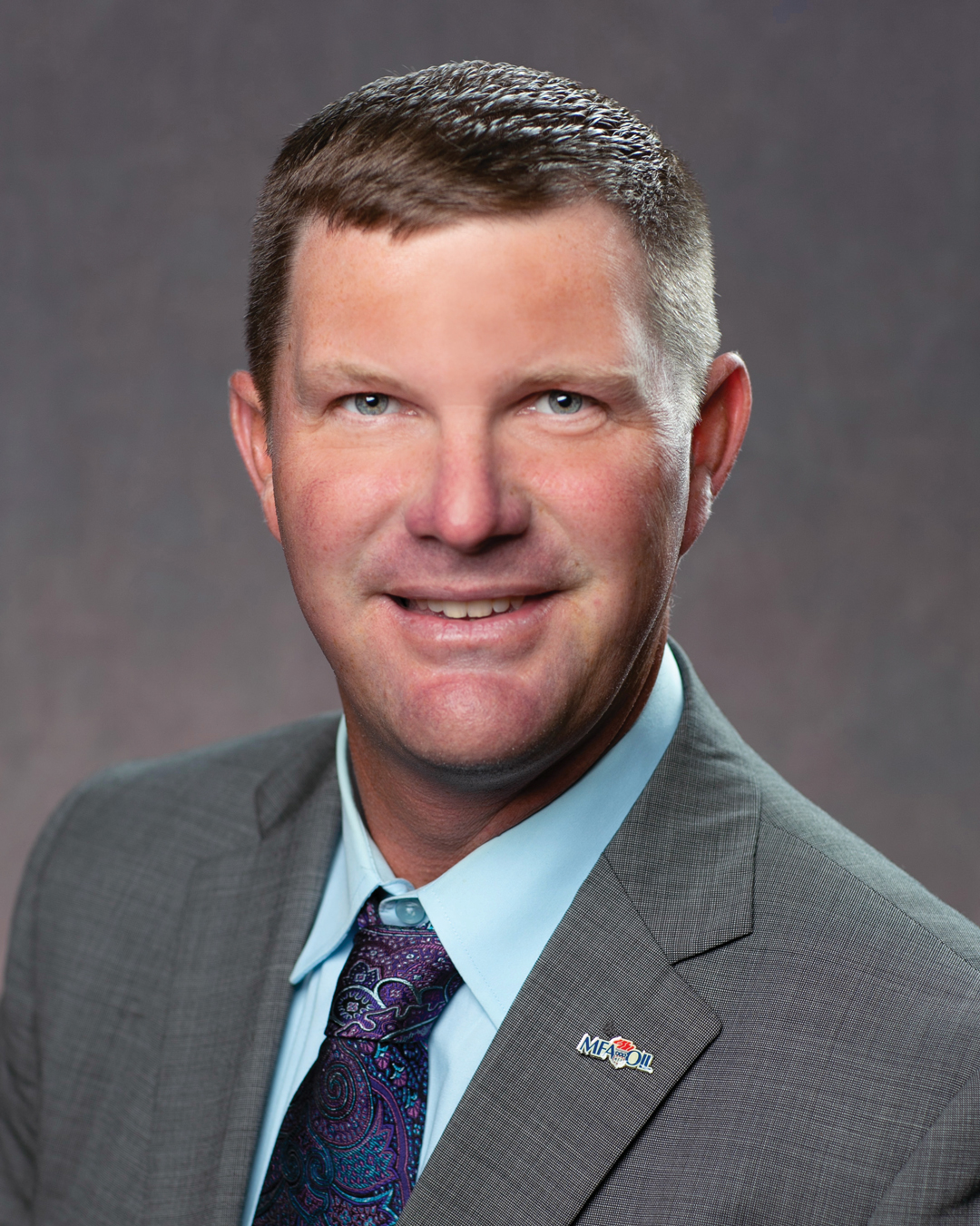 A man in a gray suit and patterned tie smiles at the camera. He has short brown hair and wears a Midco Oil pin on his lapel. The background is a plain, blurred gray.