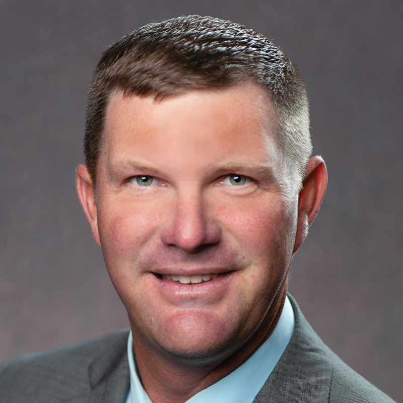 A man with short brown hair wearing a gray suit jacket, blue shirt, and no tie, smiling in front of a brownish-gray studio background.