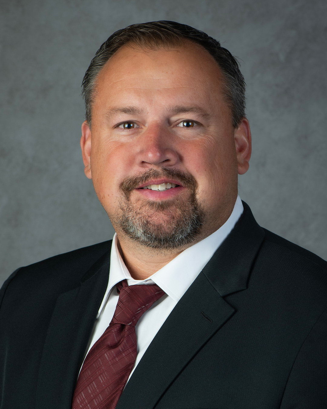 A man in a dark suit, white shirt, and maroon tie poses for a professional headshot against a gray background. He has short brown hair, a trimmed beard, and is smiling slightly.
