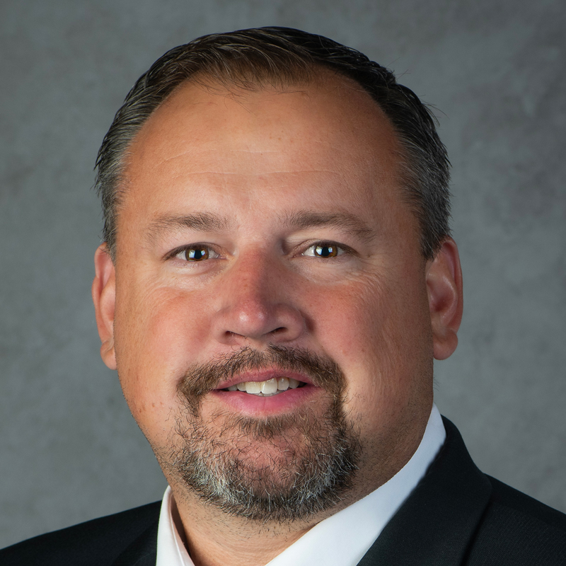 A man with short brown hair and a goatee, wearing a dark suit jacket and white shirt, smiles slightly in front of a gray background.