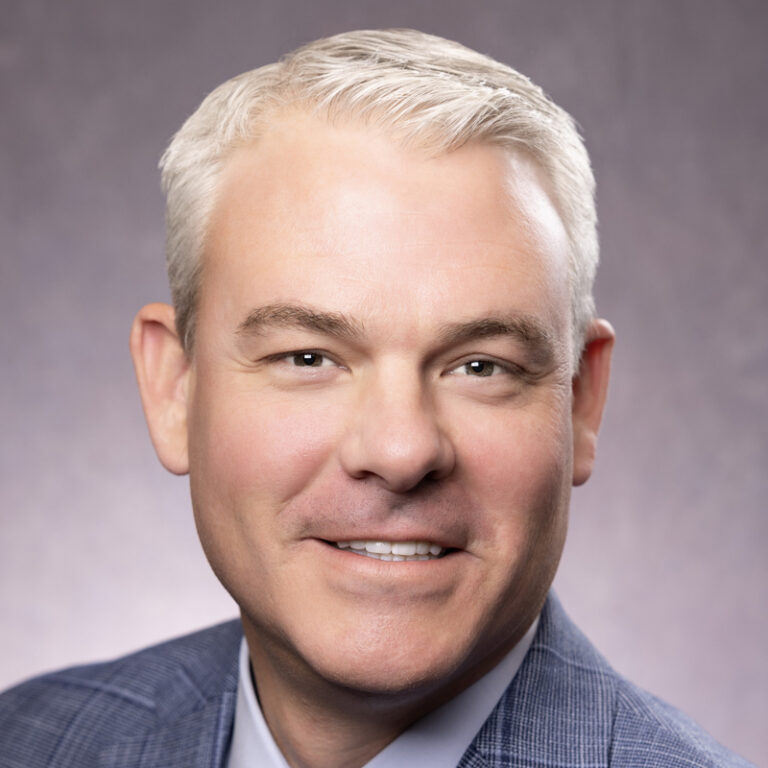 Smiling man with short gray hair wearing a blue checked suit jacket and light blue shirt, posing in front of a neutral gray background.
