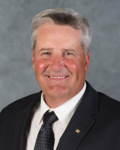 A middle-aged man with gray hair wearing a dark suit, white shirt, and patterned tie, smiling in front of a gray background.