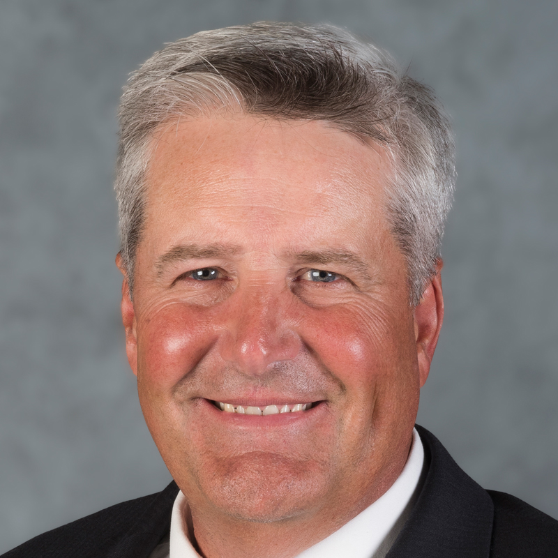 A middle-aged man with short gray hair smiles at the camera. He is wearing a dark suit jacket, white shirt, and tie. The background is a plain, muted gray.