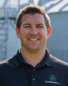 A man smiles outdoors wearing a black polo shirt with the Continuum Ag logo. Behind him are industrial metal structures and a blue sky.