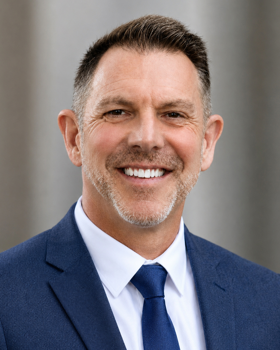 Smiling man with short brown hair and light beard, wearing a blue suit, white shirt, and blue tie, standing against a neutral blurred background.