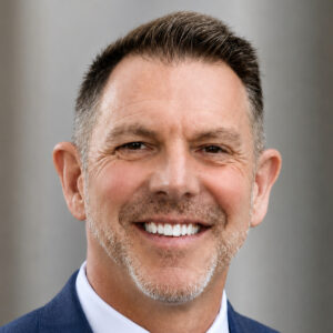 Smiling middle-aged man with short brown hair and light stubble, wearing a blue suit and white shirt, standing in front of a neutral gray background.