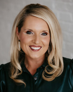 Smiling woman with long blonde hair wearing a dark green top and large earrings, posing in front of a light-colored brick wall.