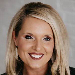 A woman with straight blonde hair, wearing large oval earrings and a dark top, smiles in front of a light-colored, blurred background.