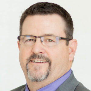 A man with short dark hair, glasses, a trimmed goatee, and mustache, wearing a gray suit and purple shirt, smiling slightly against a plain white background.
