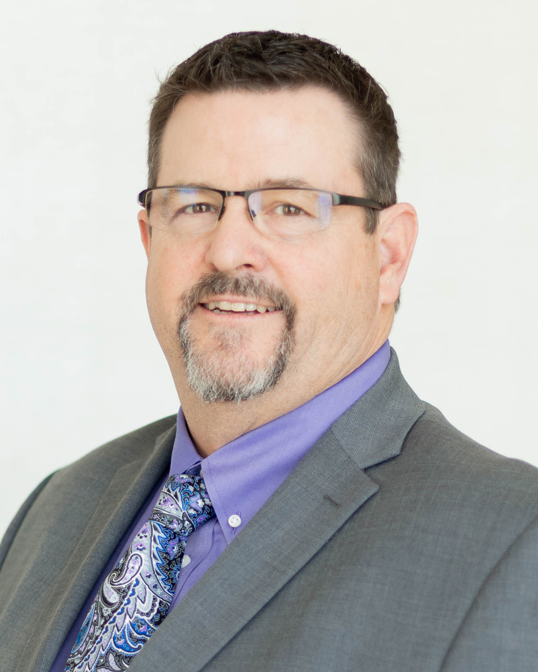 A man with short dark hair, glasses, and a goatee, wearing a gray suit, lavender shirt, and paisley tie, poses against a plain light background.